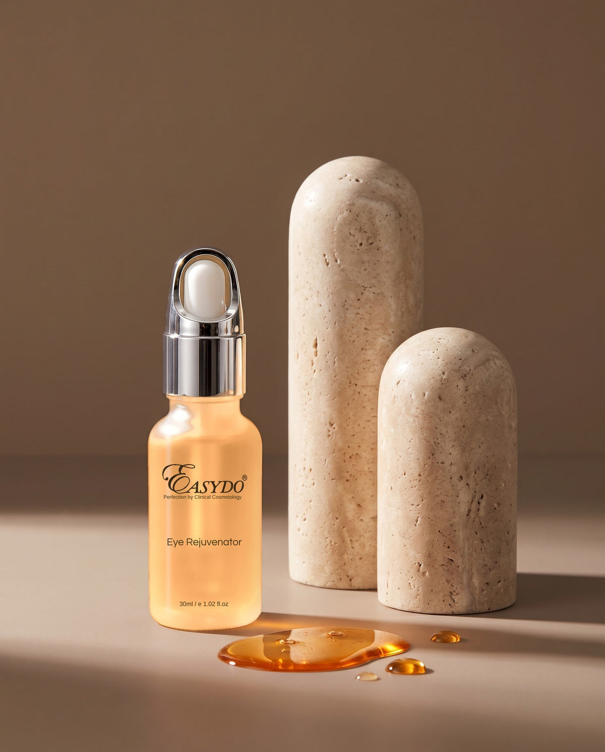 A bottle of Easynoo Eye Rejuvenator serum with a silver dropper cap, filled with golden-yellow liquid, displayed next to two textured beige stone props and a small pool of the serum on a warm beige background.