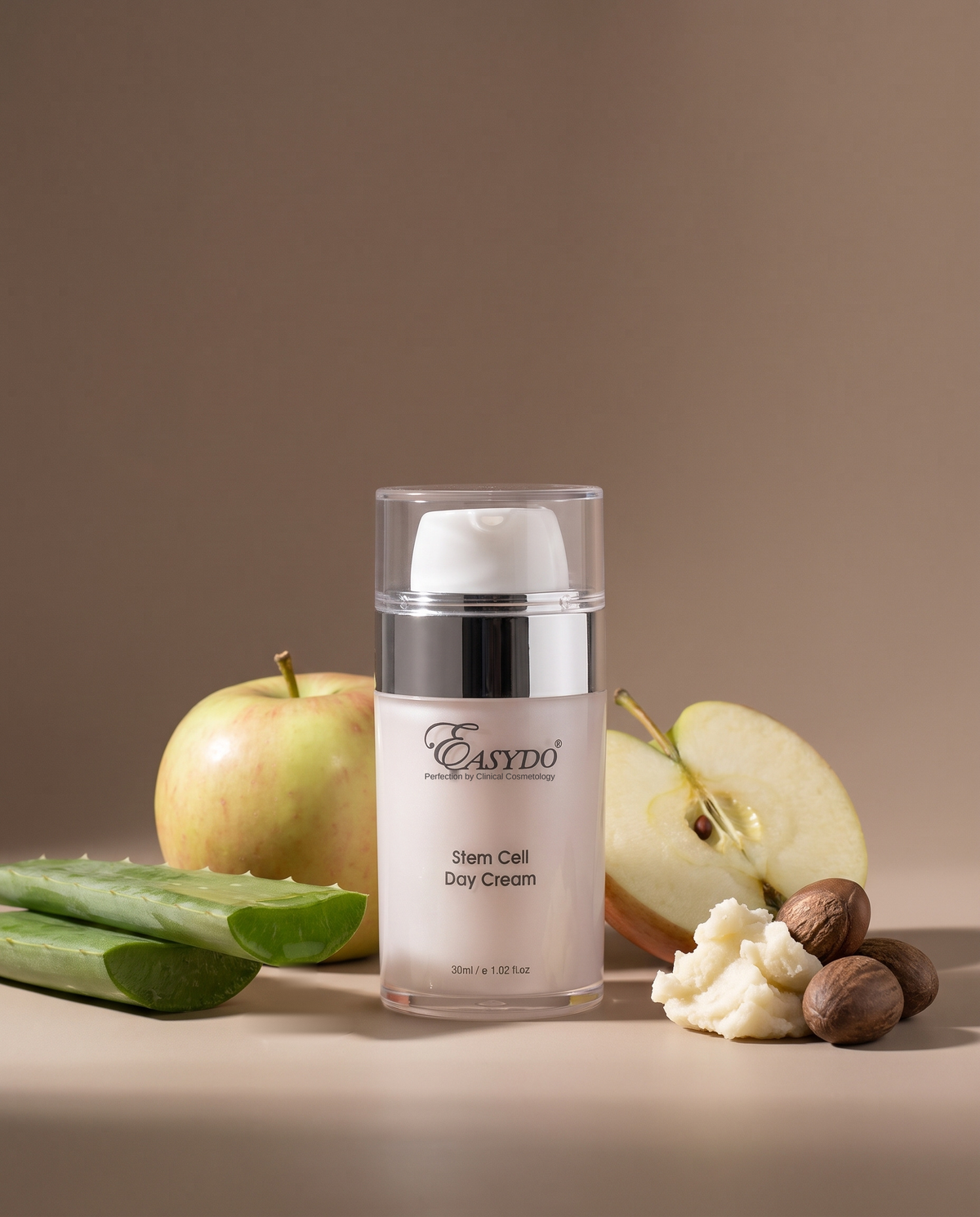 A bottle of Easydo Stem Cell Day Cream stands centered on a beige surface, surrounded by natural ingredients including a green apple, sliced apple, aloe vera, and nutmegs. The product is a clear bottle with a white pump and a silver collar, set against a solid warm brown background.